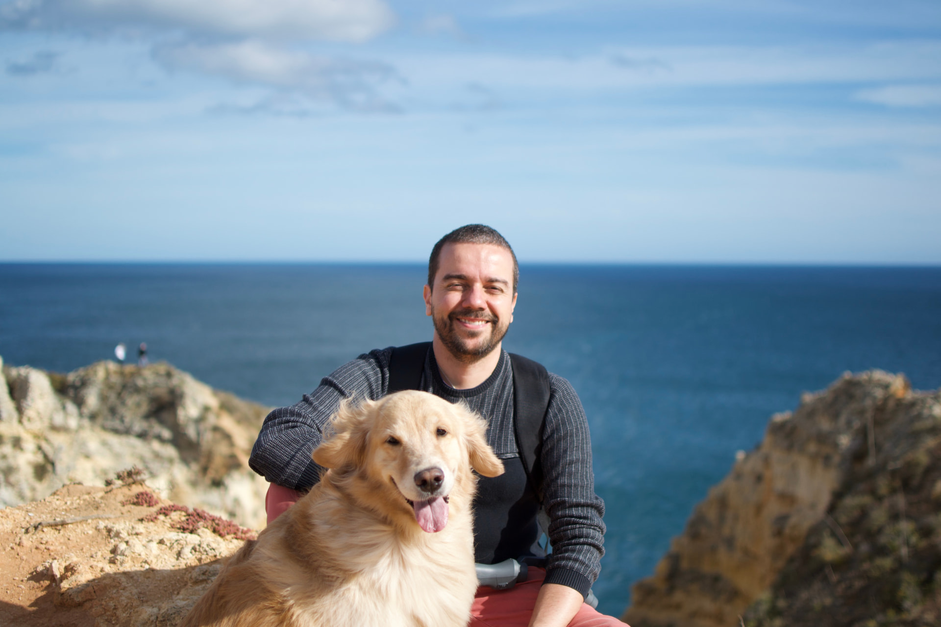 Leandro Sosi sorrindo abraçado à Julieta, sua Golden Retriever, em um mirante à beira-mar